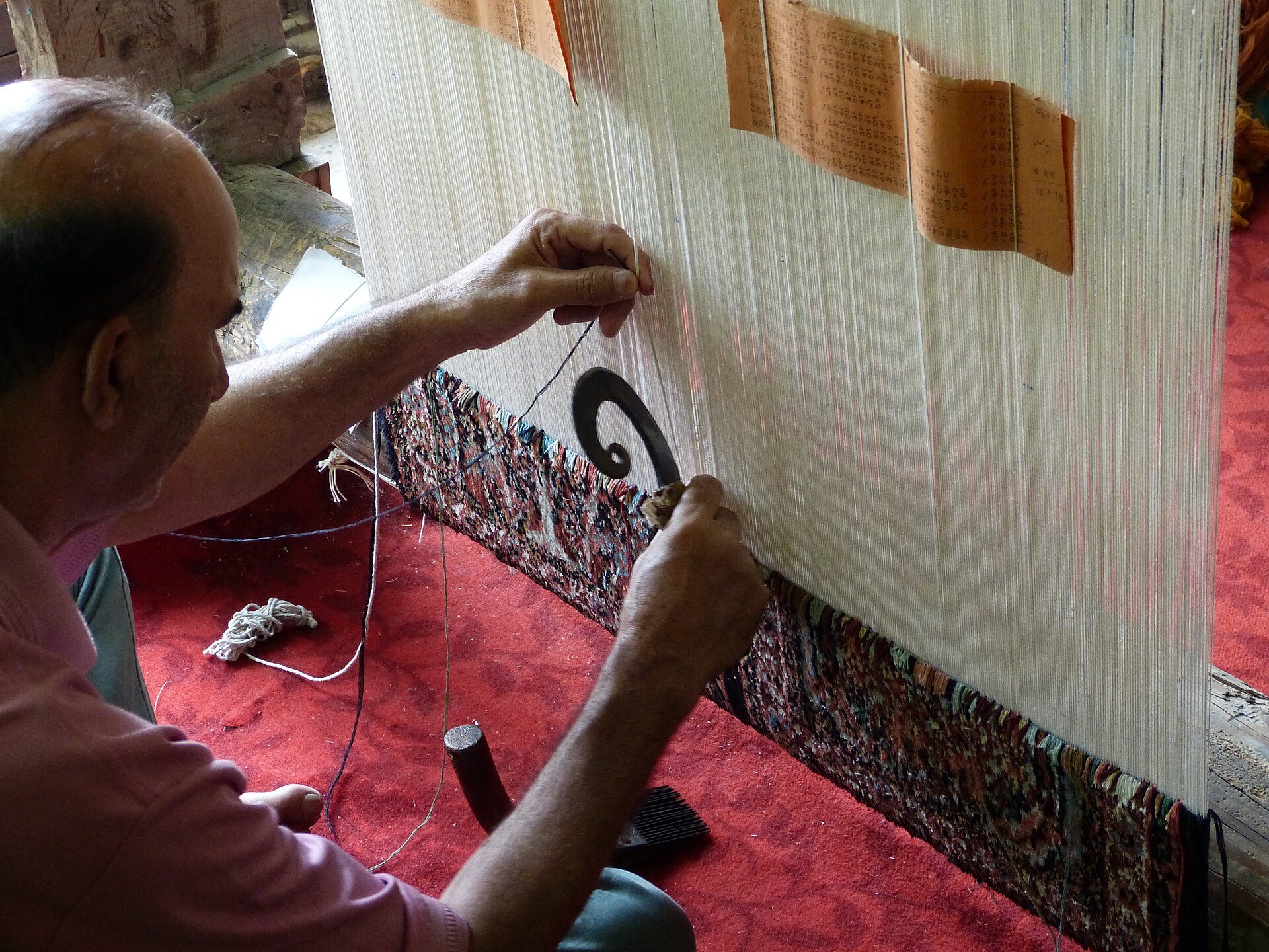 A Kashmiri weaver knotting a silk carpet, the coded talim pattern sheets pinned to the warp above his hands
