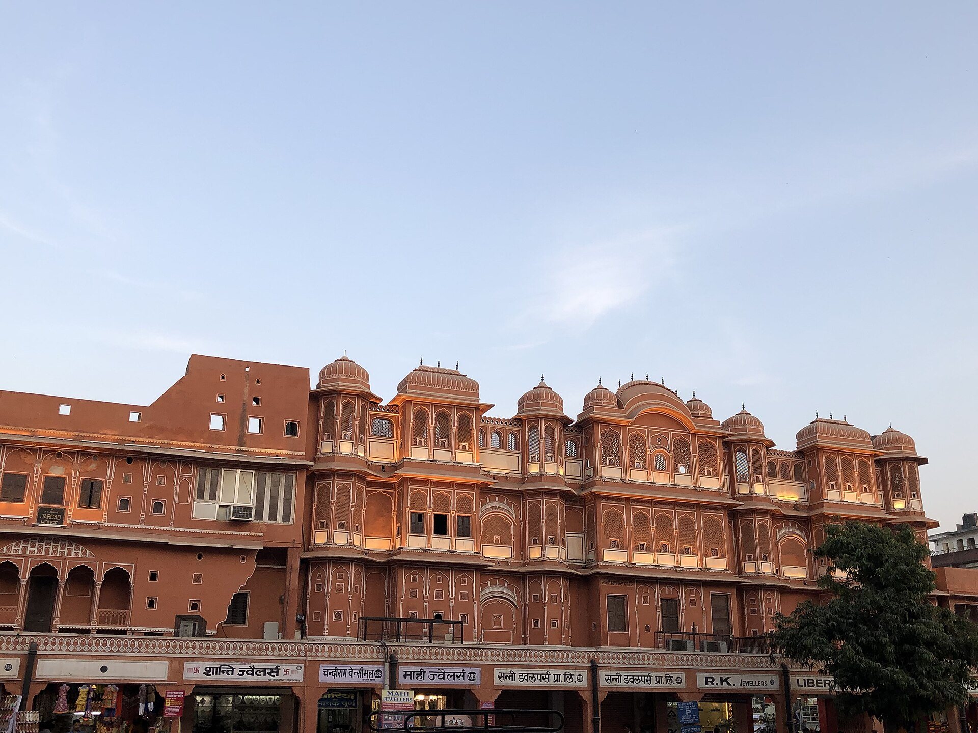 A bazaar facade in Jaipur's walled city at dusk: ornamented pink upper storeys with domed oriels above a row of jewellers' shopfronts, with an air-conditioning unit and a patched wall among them