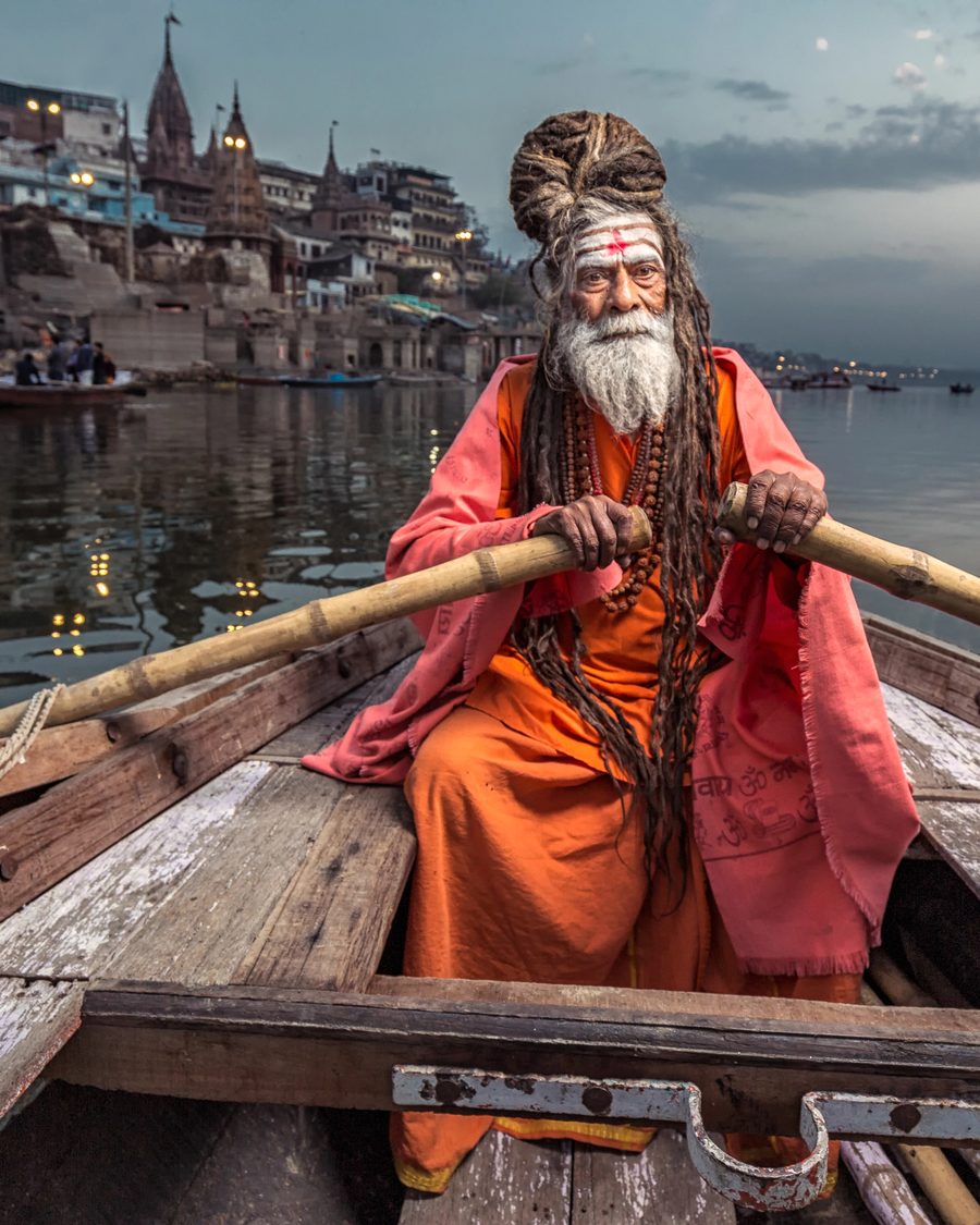 A sadhu rowing a wooden boat across the Ganges at dawn, the Varanasi ghats behind, seen from the passenger's seat